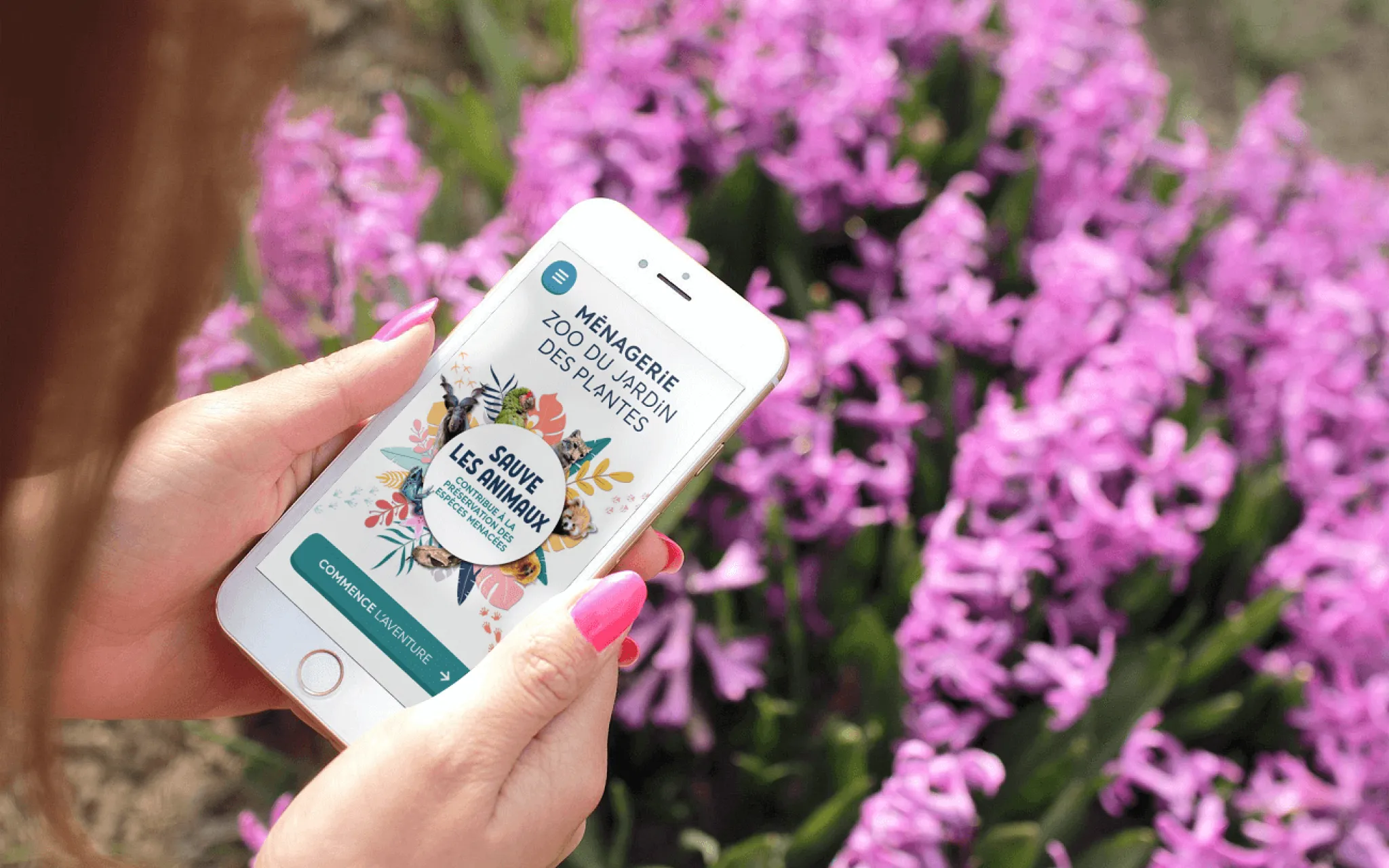 Photo montrant une femme tenant un smartphone où s'affiche la Web app de la Ménagerie, avec un massif de fleurs en arrière-plan